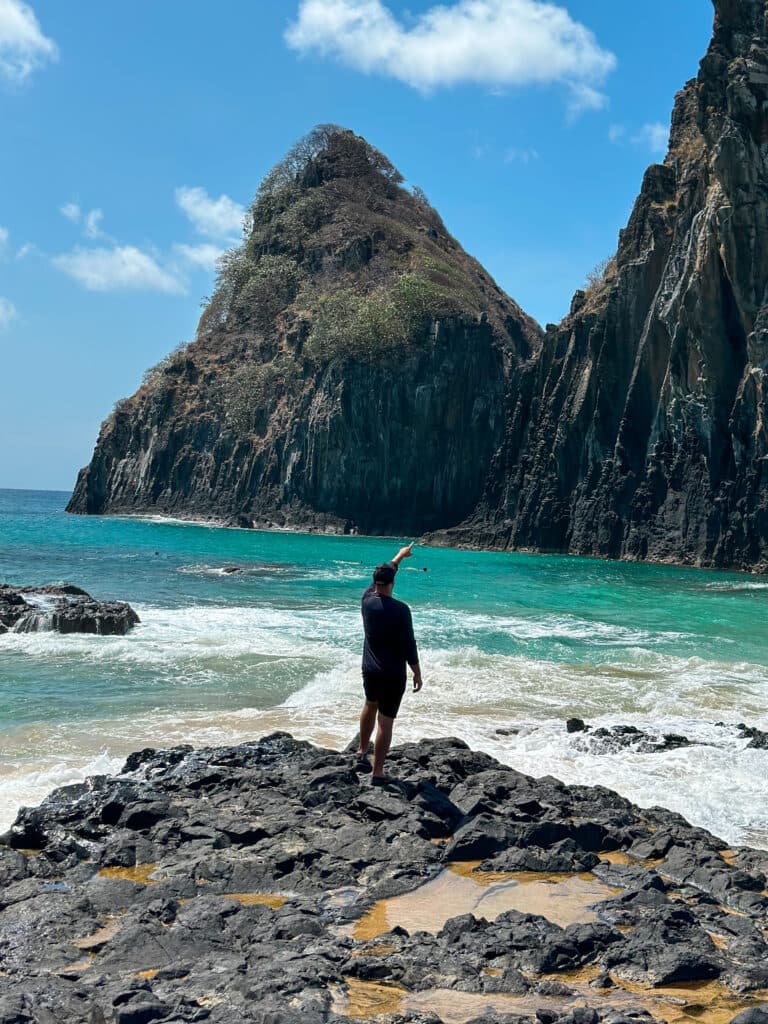 Homem em Fernando de Noronha, sobre uma pedra, olhando para o mar azul e duas montanhas