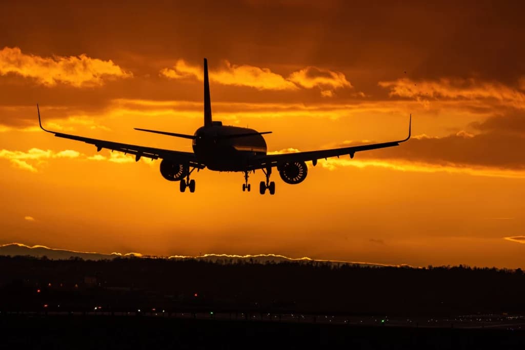 Silhueta de Avião voando com o trem de pouso aberto pronto para pousar depois de um voo na madrugada em um céu dourado num amanhecer com poucas nuvens e raios de sol